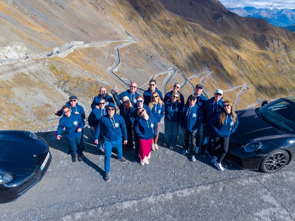 Grupo da ROXMOTORS ao lado dos Porsche no alto de um passo alpino, com as curvas da estrada ao fundo